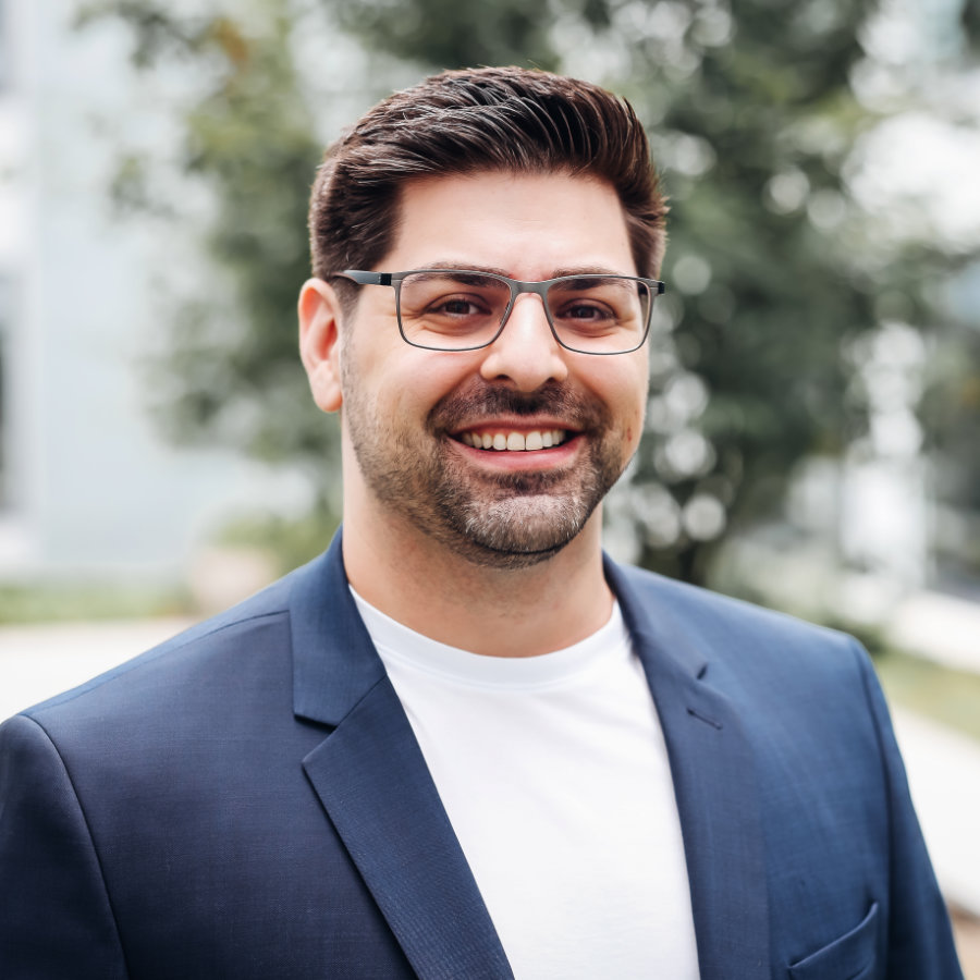 Porträt des Geschäftsführers eines IT-Unternehmens, professionell gekleidet in einem blauen Anzug. Er steht draußen und lächelt selbstbewusst in die Kamera. Als Führungskraft verantwortet er die strategische Entwicklung und digitale Transformation der Firma.