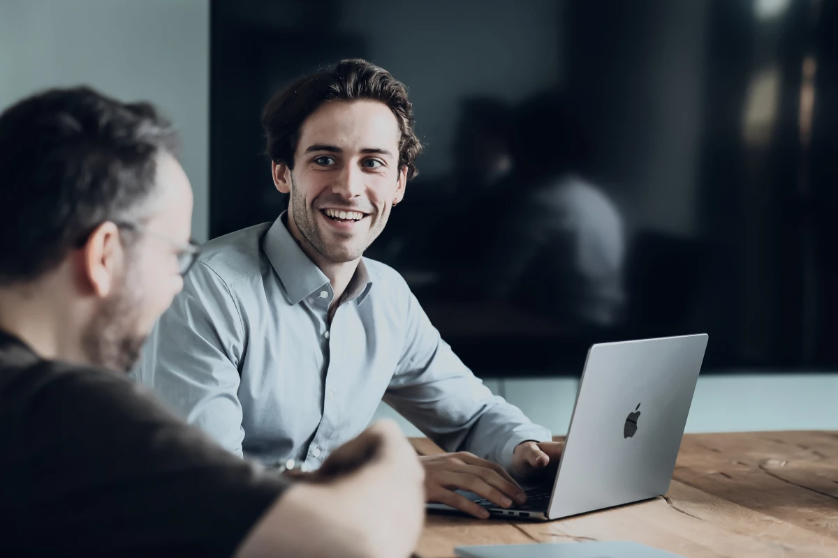 Zwei Personen im Gespräch an einem Besprechungstisch, eine arbeitet am Laptop.